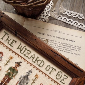 Peut inclure: Une broderie au point de croix encadr&eacute;e repr&eacute;sentant les personnages du Magicien d'Oz. Les personnages incluent Dorothy, l'&Eacute;pouvantail, l'Homme de Fer-Blanc et le Lion peureux. La broderie est sur un fond blanc avec des accents verts, bruns et rouges. Le texte "The Wizard of Oz" est brod&eacute; en fil vert.