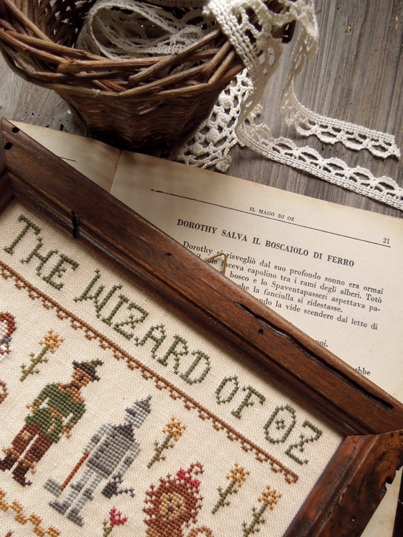 Peut inclure: Une broderie au point de croix encadr&eacute;e repr&eacute;sentant les personnages du Magicien d'Oz. Les personnages incluent Dorothy, l'&Eacute;pouvantail, l'Homme de Fer-Blanc et le Lion peureux. La broderie est sur un fond blanc avec des accents verts, bruns et rouges. Le texte "The Wizard of Oz" est brod&eacute; en fil vert.