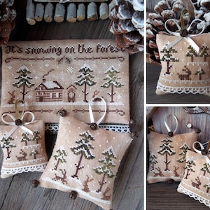 Puede incluir: Tres adornos hechos a mano con punto de cruz con un tema de bosque de invierno. Los adornos son beige con bordados marrones, verdes y blancos. Cada adorno presenta una caba&ntilde;a, &aacute;rboles y ciervos. Los adornos est&aacute;n decorados con un borde de encaje blanco y campanas marrones.