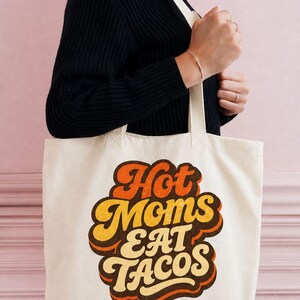 May include: A cream-colored canvas tote bag featuring the text "Hot Moms EAT TACOS" in a retro-style, layered font. The lettering uses a color palette of orange, yellow, and dark brown, with a distressed texture applied to the graphic design.