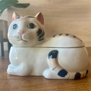 May include: A white ceramic cat-shaped lidded container featuring hand-painted dark blue tabby stripes on the head, back, and curled tail. The cat has a pink nose, green eyes, and pink-tinted inner ears, with a smooth, glossy finish.