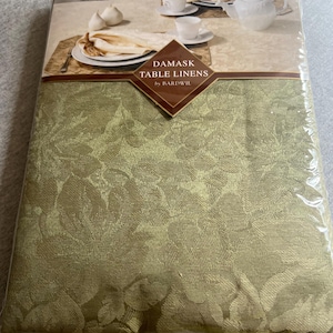 May include: A sage green damask tablecloth packaged in clear plastic. The packaging features a label with the text DAMASK TABLE LINENS by BARDWIL and an image of a table setting with a teapot, cups, and plates on a matching tablecloth.
