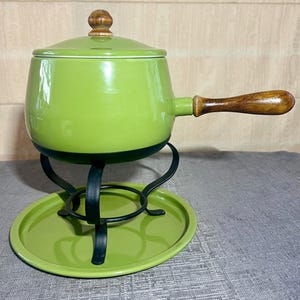 May include: A lime green enameled fondue pot with a matching lid featuring a wooden knob. The pot has a long wooden handle and rests on a black metal stand with a circular base, which is set upon a matching lime green circular metal tray.