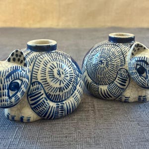 May include: Two ceramic cat-shaped candle holders featuring a cream-colored base with a blue crackle glaze finish. Each cat figure is decorated with intricate blue line patterns, including swirls and stripes, and has a circular opening on the back.