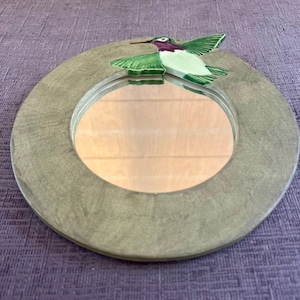 May include: A round wall mirror with a textured, muted olive-green frame. A ceramic hummingbird figurine with green wings and a purple head is attached to the top edge of the frame, extending slightly over the circular reflective glass surface.