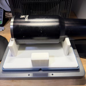 Puede incluir: Un vaso Stanley negro colocado horizontalmente sobre un soporte blanco impreso en 3D. El soporte cuenta con dos soportes laterales verticales y un bloque rectangular central para estabilizar el recipiente cilíndrico. El logotipo de Stanley es visible en el lateral.