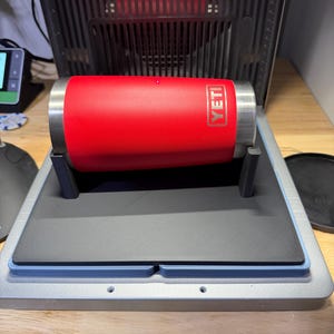 May include: A bright red cylindrical YETI tumbler with a metallic silver rim and base, positioned horizontally on a black foam support stand within a laser engraving machine. The YETI logo is clearly visible in white text on the red surface.