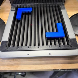 Puede incluir: Una base de grabado láser de metal plateado y negro con una rejilla de listones verticales. Dos guías de esquina de plástico azul brillante en forma de L están colocadas sobre los listones para asegurar materiales. La base incluye un marco negro.