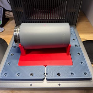 May include: A matte grey cylindrical tumbler with a metallic rim rests horizontally on a bright red plastic cradle. The cradle is positioned on a grey, multi-part base with visible screws, set within a larger machine featuring a black slatted vent.