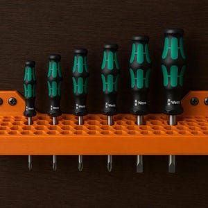May include: A set of six Wera screwdrivers with black and green ergonomic handles, arranged in a bright orange, honeycomb-patterned wall-mounted storage rack. The rack is secured with four black screws, holding the tools in vertical alignment.