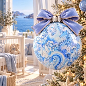 May include: A spherical Christmas ornament featuring a blue and white marbled watercolor pattern, topped with a light blue velvet bow and a silver cap, hanging from a silver cord against a blurred coastal nursery background.