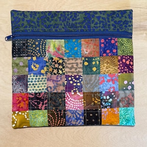 May include: A patchwork zippered pouch with a vibrant array of colorful squares. The top section features a blue and green patterned fabric, while the main body showcases a quilt-like design with various textures and hues. The zipper is a matching blue.