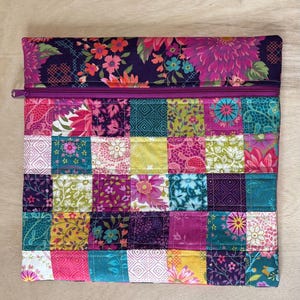 May include: A quilted patchwork zippered pouch featuring a variety of floral and geometric patterns in shades of pink, purple, teal, and yellow. The top section has a dark purple floral print, and the zipper is a matching purple color. The pouch is rectangular.