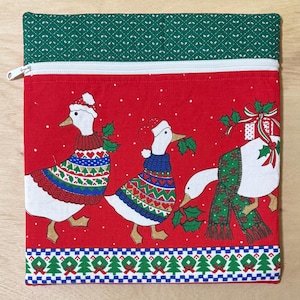 May include: A square fabric zipper pouch featuring a red background with three white geese wearing Santa hats and colorful patterned sweaters. The pouch has a green top border with a white pattern and a bottom border with a Christmas tree design.