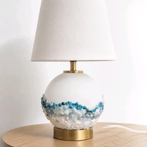 May include: A table lamp with a white conical fabric shade and a spherical white glass base. The lower half of the glass sphere is decorated with a wave-like pattern of small, polished blue and white stones. It has a brass-toned metal base.