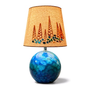 May include: A table lamp with a spherical base featuring a glossy, marbled blue and teal finish. The tapered, tan-colored fabric lampshade is decorated with a pattern of orange spiral seashells and small, scattered blue and green sea glass pieces.