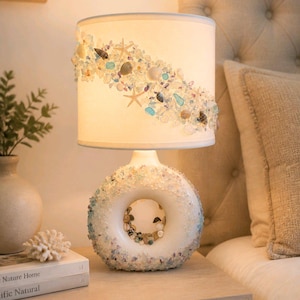 May include: A table lamp with a white cylindrical shade decorated with a diagonal band of small seashells, starfish, and blue and clear glass fragments. The base is a white, donut-shaped ceramic form covered in similar shells and glass.