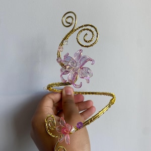 May include: A gold-coloured wire arm cuff featuring a spiral design with two translucent pink flower-shaped beads. The wire is wrapped in a coil pattern and accented with small clear and purple beads, creating a decorative piece of jewellery.