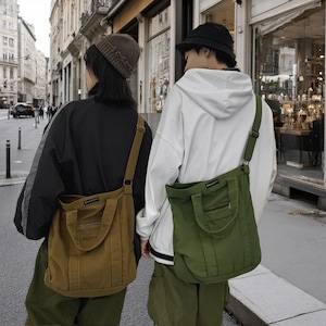 May include: Two canvas tote bags with adjustable shoulder straps and top handles. One bag is brown and the other is olive green. Each bag features a small rectangular label on the upper front and a horizontal zippered pocket below the handles.