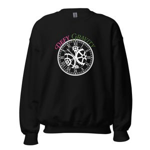 May include: A black long-sleeve sweatshirt featuring a white graphic of a clock face with visible internal gears. Above the clock, the text "DEFY GRAVITY" is printed in a curved arc, with "DEFY" in pink and "GRAVITY" in green lettering.