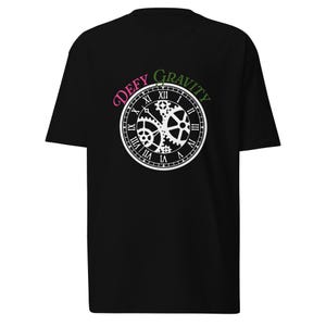 May include: A black short-sleeved t-shirt featuring a white graphic of a clock face with visible internal gears and Roman numerals. The text 'DEFY' is printed in pink and 'GRAVITY' is printed in green, arching over the top of the clock design.