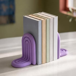 Può includere: Una coppia di fermalibri viola con un design ad arco scanalato che sostiene una fila di cinque libri. I libri hanno copertine a tinta unita nei toni del grigio chiaro, beige, verde pallido e bianco, posizionati verticalmente tra i due supporti.