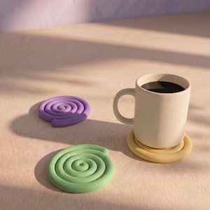 May include: A white ceramic mug filled with dark coffee resting on a yellow spiral coaster, accompanied by a light green spiral coaster and a purple spiral coaster. Each coaster features a continuous coiled design made of a smooth, solid material.