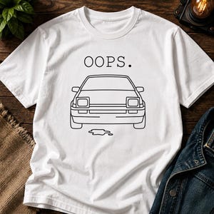 May include: A white short-sleeve t-shirt featuring a black line art illustration of the front of a car with pop-up headlights. Above the car, the word OOPS. is printed in black capital letters, and a small puddle graphic is below the car.