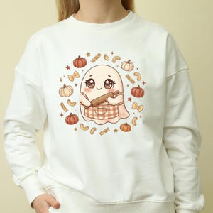 May include: A cream-colored crewneck sweatshirt featuring a central graphic of a cute cartoon ghost wearing a checkered apron and holding a wooden rolling pin, surrounded by a circular pattern of small orange pumpkins, pasta shapes, and stars.