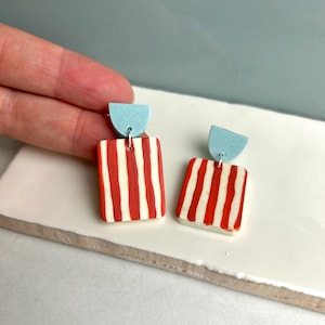 Puede incluir: Un par de pendientes hechos a mano. Cada pendiente presenta una parte superior de semicírculo azul claro y una parte inferior rectangular con rayas verticales rojas y blancas. Los pendientes se exhiben sobre una superficie blanca.