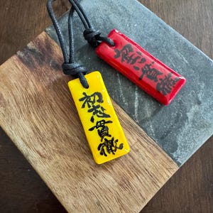 Könnte beinhalten: Zwei rechteckige Anhänger-Halsketten mit schwarzen Kordeln. Ein Anhänger ist gelb mit schwarzen japanischen Schriftzeichen, der andere ist rot mit schwarzen japanischen Schriftzeichen. Die Anhänger liegen auf einer Holz- und Steinoberfläche.