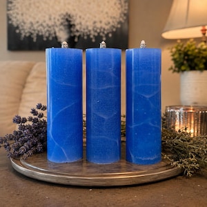 May include: Three cylindrical blue candles with a visible crackle texture and white veining patterns. Each candle features a small silver-colored metal wick holder at the top, standing upright on a flat, circular, metallic-toned base.