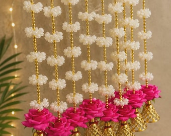 May include: A decorative hanging garland featuring multiple vertical strands of small white jasmine-like flowers interspersed with gold beads. Each strand terminates in a vibrant hot pink rose bloom attached to a gold-toned, bell-shaped metal lattice.