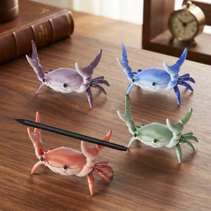 Peut inclure: Quatre petites figurines en forme de crabe de couleurs violette, bleue, verte et corail. Chaque crabe possède des pinces relevées conçues pour tenir un stylo ou un crayon. Un stylo noir repose horizontalement sur les pinces du crabe corail.