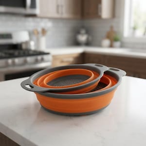 May include: A set of two round, collapsible kitchen colanders featuring bright orange silicone sides and dark grey plastic rims and bases. Each colander has integrated handles on opposite sides and a perforated bottom for draining liquids.