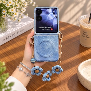 May include: A blue denim-textured foldable smartphone case featuring a circular magnetic ring on the back and a decorative strap adorned with blue fabric flowers and a striped bead. The phone screen displays the text Xiaomi MIX Flip.