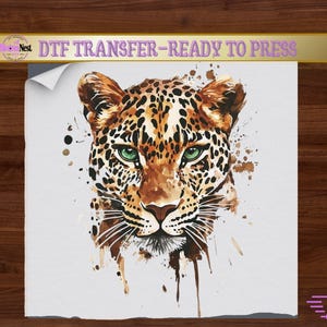 May include: A DTF transfer ready to press featuring a watercolour-style illustration of a leopard head with striking green eyes, black spots, and brown fur tones. The design includes paint splatter effects on a white background with text at the top.