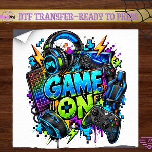 Peut inclure: Un transfert DTF avec le texte GAME ON en bleu et vert, entouré d'équipements de jeu incluant un casque, un clavier, une souris, deux manettes et une chaise de jeu, le tout agrémenté d'éclaboussures de pixels aux couleurs vives.