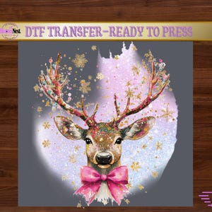 May include: A DTF transfer ready to press featuring a deer head illustration with a pink bow, gold snowflakes, and colorful paint splatters against a light purple and white circular background. The text reads DTF TRANSFER-READY TO PRESS.