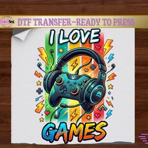 May include: A DTF transfer sheet featuring a colorful graphic of a black gaming controller wearing large headphones. The design includes the text I LOVE GAMES, surrounded by vibrant vertical stripes, yellow lightning bolts, small stars, and hearts.