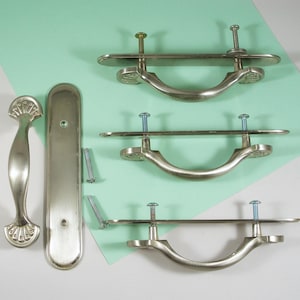 May include: A collection of brushed nickel cabinet hardware, including three drawer pulls, a door handle, and a long backplate. The drawer pulls have a curved handle design with decorative end details. Screws are included.