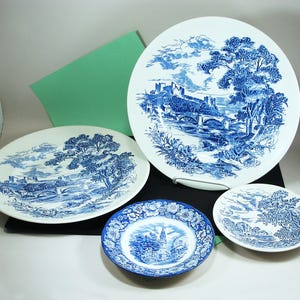 May include: A collection of four white ceramic plates featuring blue transferware patterns. The designs depict detailed landscapes with castles, bridges, trees, and a church steeple surrounded by intricate floral borders on the smaller plates.