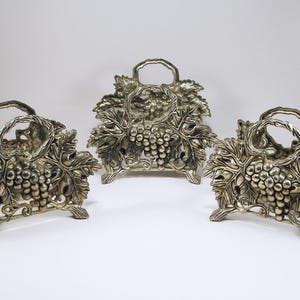 May include: Three silver-colored napkin holders with a grape and leaf design. Each holder has a handle and is shaped like a basket. The holders are made of metal and are on a white background.
