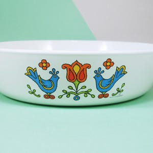 May include: White ceramic bowl with a colorful bird and floral design. The bowl features two blue birds with yellow accents, orange flowers, and a central orange and yellow flower. The text "1975 Corning" is visible.