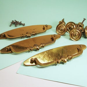 May include: Three brass drawer pulls with a curved handle and ornate detailing.  Five brass knobs with a raised center and a decorative edge.