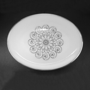 May include: Oval white serving platter with a silver rim. The center features a black, symmetrical floral design. The platter is set against a dark background, highlighting its elegant design.