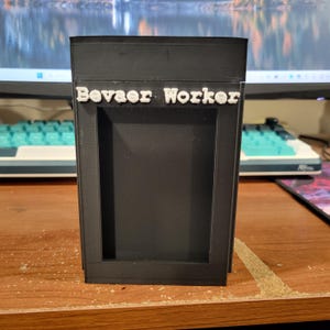 May include: A black, rectangular, 3D-printed container featuring a hollow center and the white, raised text "Bevaer Worker" positioned horizontally across the upper front section. The object has a smooth, matte finish and a vertical orientation.