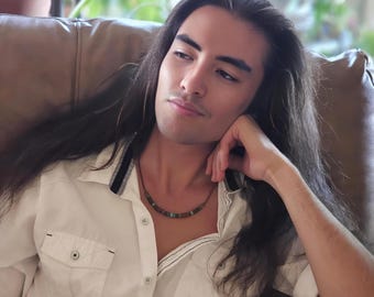 May include: A person with long dark hair is sitting in a chair. They are wearing a white button-down shirt with a beaded necklace. The person is looking off to the side.