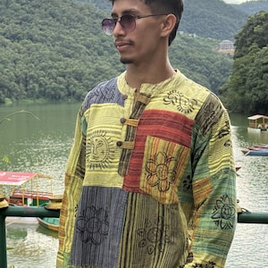Pode incluir: Uma camisa de manga comprida com um design de patchwork com vários painéis em tons de amarelo, verde, vermelho e cinzento. O tecido apresenta motivos pretos estampados de sóis e flores. A camisa tem uma gola mandarim e três botões de madeira.