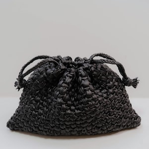 Pode incluir: Uma bolsa com cordão preta feita de um material tecido espesso. A bolsa apresenta um padrão texturizado e entrelaçado em todo o corpo e é fixada no topo com dois cordões pretos trançados que se puxam para fechar a abertura da bolsa.